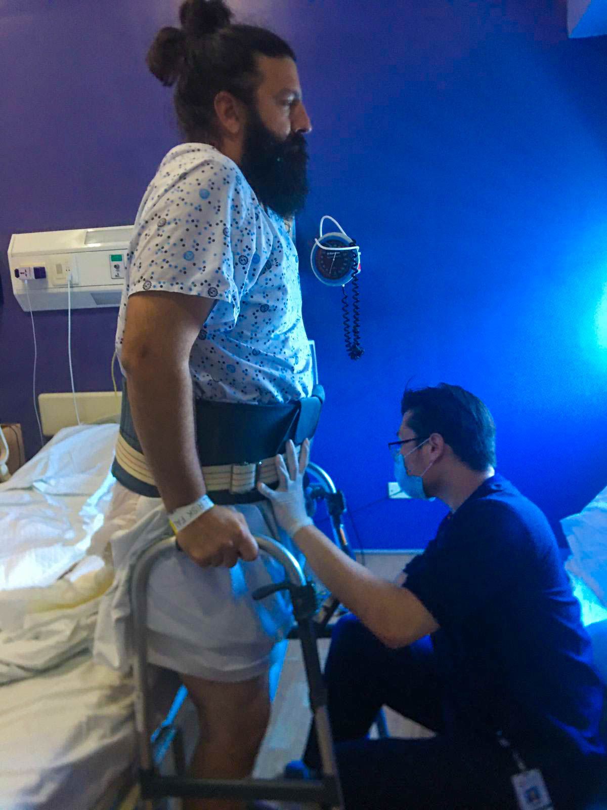 Photo of Chris standing with help from a walker and physical therapist on the first day after his disc removal surgery.